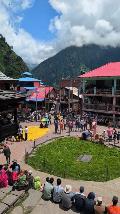 The image depicts a vibrant community gathering in the village of Malana, India, likely during a local festival or event. The scene is set in an open courtyard surrounded by traditional multi-story buildings with colorful roofs, predominantly red and blue. The architecture suggests a mountainous region, with lush green hills and pine forests visible in the background, under a partly cloudy sky.
In the foreground, a group of people are seated on stone steps, facing away from the camera, watching the activities in the courtyard. They are dressed in a mix of traditional and modern attire.
The courtyard itself is bustling with activity. A large, bright yellow tarp is spread out on the ground, with several individuals standing or sitting around it. Many other people are milling about, some in small groups engaged in conversation, while others appear to be observing the event. There are also some children present, adding to the lively atmosphere.
Several buildings in the background feature balconies where more people are gathered, suggesting a communal viewing area or part of the celebration. The overall impression is one of a well-attended, traditional gathering in a scenic, rural setting.