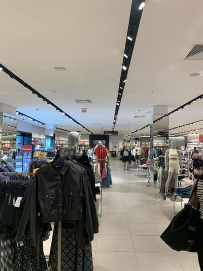 This image presents the interior of a brightly lit retail clothing store, identified as a Riachuelo department store located in Camaragibe, Brazil. The scene is characterized by a high white ceiling equipped with black track lighting fixtures and light-colored square floor tiles.
In the immediate foreground, a black faux leather-style jacket is displayed on a clothing rack, with a black and white patterned skirt visible beneath it. To the left, other clothing racks hold an assortment of dark-colored garments, with a visible sign displaying "POOL JEANS" and a yellow price tag indicating "R$ 29."
The central and mid-ground sections reveal a wide aisle where several mannequins are positioned, showcasing various outfits, including a light-colored crop top and shorts ensemble. On the right side of the mid-ground, a person is partially visible from the mid-thigh down, walking away from the camera and carrying a large black shopping bag with the word "RIACHUELO" printed in white.
The background shows further sections of the store with more clothing displays and shelving units. A red "SAÍDA" (Exit) sign is visible high on a wall in the central background, along with a directional sign partially displaying Portuguese text, including "PROVADORES" (Fitting Rooms) and "CAIXAS" (Checkouts). Another individual is discernible walking in the distant background. The overall environment is consistent with a commercial apparel retail space designed for customer browsing and shopping.
