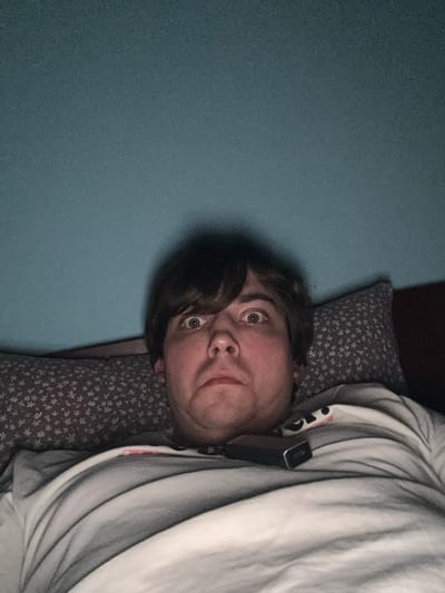 A person is lying in bed, facing the camera. The person has light brown hair and is looking up with wide eyes, a surprised expression. They are wearing a grey and white striped t-shirt. A black object, possibly a vape device, is resting on their shirt. The background consists of a teal wall and two pillows, one with a white floral pattern on a grey background, and the other a darker color. The lighting is dim, creating a shadow behind the person's head. The image appears to have been taken in a bedroom.