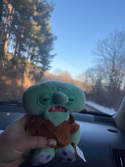 The image displays a mint green plush toy, resembling "Handsome Squidward," held by a person's hand inside a vehicle. The toy, approximately 15-20 cm in height, wears a brown shirt and features embroidered tired eyes, a prominent nose, and an open mouth revealing a pink interior and two white teeth; purple polka-dot patterns are visible on its feet, and a white tag hangs from its left leg. The hand holding the toy exhibits manicured nails with a light, shimmering polish.
The background, viewed through the vehicle's front windshield, shows an outdoor environment consistent with the Ichera, Bulgaria area. To the left, a dense forest of sunlit coniferous trees is partially obscured by bokeh. To the right, bare deciduous trees are visible against a clear blue sky. A dark paved road, with traces of snow or light debris on its right edge, recedes into the background. Portions of the vehicle's dark gray dashboard and an air vent are visible in the lower right corner of the frame. The scene is illuminated by bright natural sunlight, suggesting a clear day during winter or late autumn.