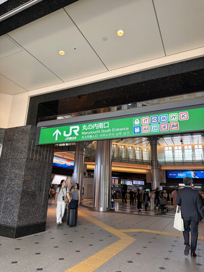 The image depicts the Marunouchi South Entrance of Tokyo Station in Chiyoda, Japan. A large green sign with the JR East logo and text in Japanese, English, and Korean announces the "Marunouchi South Entrance." Below the main sign, there are rows of colored squares, some containing letters and symbols.

In the foreground, two individuals are walking with luggage. The person on the left, wearing white pants and a grey top, is pulling a black suitcase. The person on the right, in a patterned top and dark pants, is also accompanied by a suitcase.

Further into the station, several other people can be seen, including families with children and individuals dressed in business attire. There are also information boards and digital displays visible. The station architecture features tall pillars and a brightly lit interior with large windows. The flooring is a patterned tile with yellow tactile paving for accessibility. The overall impression is of a busy, modern transportation hub.