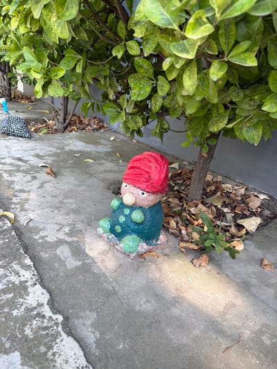 A gnome statue with a red hat and teal body sits on a concrete walkway next to a bush in Balakənd, Azerbaijan. A guinea fowl statue is visible to the left of the gnome. The ground is covered in fallen leaves.