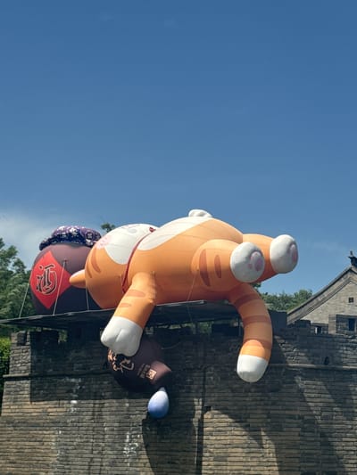 An upward-looking shot captures a large, inflatable orange tabby cat with white paws. The cat is suspended in the air near an old, gray brick wall and against a bright blue sky. At the bottom of the cat, there is a dark balloon with Chinese lettering, possibly indicating a brand or location. The scene is set near green trees, some of which are visible in the background. The location is the city of Miyun District, China.