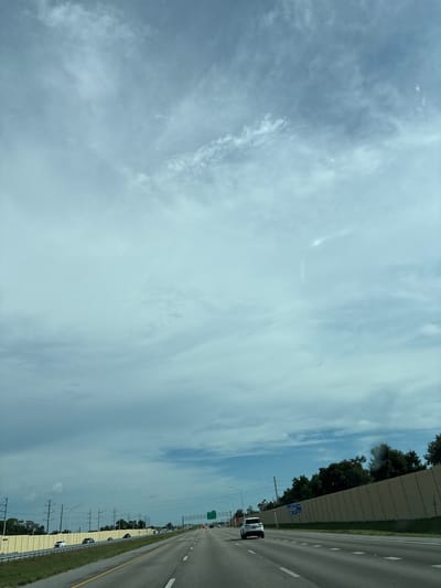 The image depicts a highway scene on a cloudy day. A multi-lane road stretches into the distance, with several vehicles on it. On the right side of the road, a tall, beige sound barrier runs along the perimeter of a wooded area. On the left side, there's a similar sound barrier, and beyond it, utility poles and wires are visible. The sky is filled with a mix of light and dark clouds, creating a diffused lighting effect. The location is identified as being in Sarasota, United States.