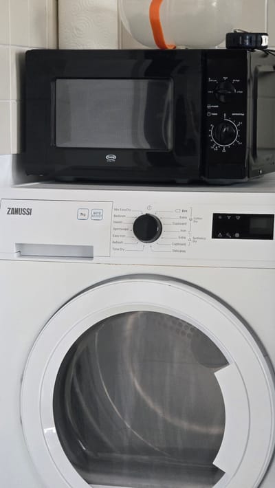 This is a vertical, eye-level, medium shot photograph of a black microwave oven stacked on top of a white Zanussi washing machine. The microwave is positioned centrally at the top of the frame, and the washing machine fills the bottom two-thirds of the frame. The microwave has a dark glass door and a control panel with a circular dial and buttons on the right side. The Zanussi washing machine has a circular door with a metallic drum visible inside. The control panel of the washing machine is located on the top right and features a large dial with various laundry program selections, such as "Mix EasyDry," "Bedlinen," "Denim," and "Delicates." Both appliances appear to be in a domestic setting, likely a kitchen or utility room. The background consists of a tiled wall and a roll of paper towels. The image is well-lit and shows the appliances from the front. There are no people or animals visible in the image. The image does not contain any information about the city of Kilkenny, Ireland.