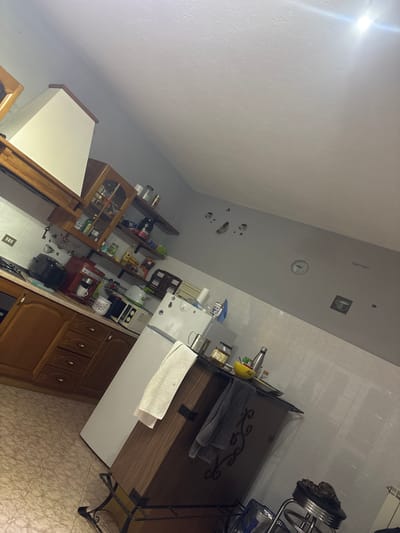 The image depicts a cluttered kitchen in San Pietro di Caridà, Italy. White walls and tiled floors surround wooden cabinetry filled with various items. Appliances, containers, and utensils cover the countertops. There are no people visible.