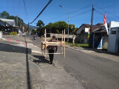 The image depicts a street scene in Gadog, Indonesia, under clear blue skies with bright sunlight. In the foreground, an individual wearing a dark cap, a plaid shirt, and dark trousers walks away from the camera on the right side of an asphalt road. This person is carrying a light-colored, rectangular wooden frame, consistent with the structure of a portable vendor's stall, on their back. The frame casts a distinct shadow on the asphalt.
To the left of the pedestrian, a concrete sidewalk with a painted red and white curb slopes downwards. Further along this left side, multiple buildings are visible, including one with a white facade and green elements displaying numerous stacks of yellow fruit. Several red and white Indonesian flags are affixed to poles and utility wires along the street.
The asphalt road extends into the distance, appearing to gently ascend. Several motorcycles are visible traveling further down the road. On the right side of the road, opposite the main pedestrian, are buildings with varying architectural styles, including one with a blue section and another with a prominent brown roof. Utility poles with multiple overhead wires span across the frame. Trees and dense vegetation are visible in the background, and distant hills or mountains are discernable under the horizon.
