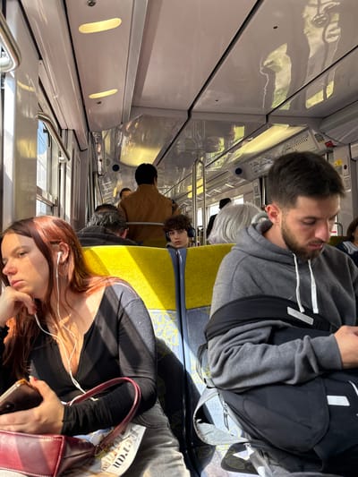 The image shows the interior of a train carriage, taken from the perspective of someone sitting on the aisle seat, facing the window. The carriage is brightly lit. Several passengers are seated, including a young woman with long red hair wearing a black top and jeans who is holding a black smartphone and a dark-colored purse. She is wearing headphones and looking down at her phone. Next to her is a young man with dark hair wearing a gray hoodie. He has a black backpack on and is looking at his phone. In the background, there are more passengers, including a young man with dark hair wearing a black and yellow jacket, and a man with a brown jacket, seated facing the front of the carriage. This train carriage is likely part of the RER B line in France, given the yellow seats and general design, and the location of the image is a train station in the city of Bourg-la-Reine, based on the provided coordinates. The environment appears to be a public transit system. The image shows a typical scene of people commuting on public transportation in France.