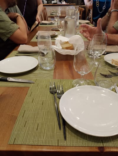 This is a close-up, eye-level, medium shot of a wooden table set for a meal at what appears to be a restaurant, with multiple place settings. The table is covered with light green woven placemats. On the left side of the frame, a white dinner plate is positioned on a placemat, with a knife placed to its left. Further to the left, a person wearing a green t-shirt and a partial view of their arm and hand are visible. They are wearing a bracelet. In the background, other people are partially visible. To the right of the plate, a clear wine glass is empty. Behind it, another clear glass is visible, with forks inside. Further to the right, another white plate with bread and a paper napkin is set. A carafe filled with water and other glasses are also visible. The overall setting suggests a communal dining experience. The restaurant is located in the city of Torri in Sabina, Italy.