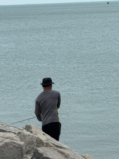 A man in a wide-brimmed hat and long-sleeved gray shirt stands on a rocky outcrop. He's wearing black pants. He is fishing, holding a rod that extends downwards. The backdrop features a vast expanse of water, with light reflecting on its surface. A dark, indistinct object is visible in the far distance over the water. The photo is rotated.