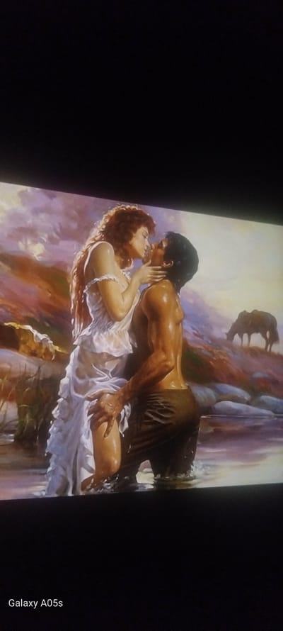 The image is a photograph of a painting, likely a romance novel cover, presented at an angle with black borders on the left, top, and bottom. The photograph includes the text "Galaxy A05s" in the bottom right corner.

The painting depicts a man and a woman in a passionate embrace within a lush, wet, and rocky natural setting. The man is on the right, muscular and bare-chested, with dark, wet hair. He wears a dark, tattered cloth around his lower body. His skin glistens with water as he leans over the woman, his right arm around her waist and his left hand near her face. The woman, on the left, has reddish-brown, curly hair and is wearing a flowing white dress or chemise, which also appears wet and clings to her body. Her left hand is raised to the man's face, while her right arm is out of view. Their faces are close, suggesting they are about to kiss or are in the midst of a kiss. Both figures appear drenched, indicating they might be in or near a waterfall or stream.

The background of the painting is rendered with broad, abstract brushstrokes in shades of purple, brown, blue, and green, evoking wet rock formations, cascading water, and foliage. A dark, indistinct shape in the upper background could be a further rock outcrop or distant trees. The overall mood is romantic and intense, focusing on the intimate moment between the two figures. There are no specific cues within the painting to indicate a location like Zavoy, Bulgaria; the setting is generalized as a wild, natural landscape.