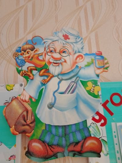 A colorful, cartoon-like cutout depicts an elderly, smiling doctor wearing glasses, a white coat, and a blue and green striped pair of pants. The doctor has white hair and a white mustache and beard. A red cross is on the doctor's blue cap. The doctor is holding a brown medical bag in their left hand. The doctor's right hand is holding a small blue bottle labeled "AB." A small monkey with an orange and yellow shirt is perched on the doctor's left shoulder, holding a blue telephone to its ear. A stethoscope is draped around the doctor's neck, with the earpiece in the doctor's chest pocket. A thermometer is also visible in the pocket. A yellow butterfly with black stripes is on the doctor's coat. The background is a light brown wallpaper with vertical, wavy lines. The cutout is affixed to a wall. The image is taken in Zhirnovsk, Russia.