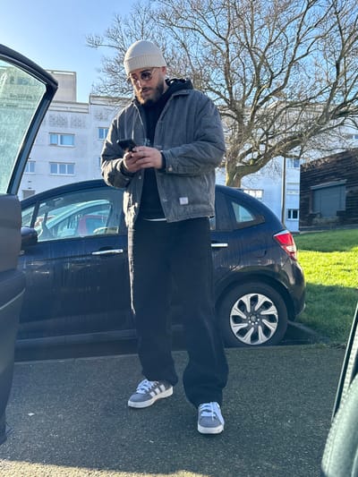 An adult male stands outdoors in Hérouville-Saint-Clair, France, beside a dark hatchback car with its front left door ajar. He holds a dark-colored smartphone horizontally with both hands, his gaze directed downwards towards the device. The male wears a light-colored beanie, round sunglasses with light frames, a dark grey quilted jacket with a white label on the lower left, a black hooded sweatshirt underneath, black trousers, and grey sneakers with white stripes and laces. A silver ring is visible on his left pinky finger. The car is parked on an asphalt surface, adjacent to a grassy area. In the background, a light-colored, multi-story residential building with multiple visible windows, a large bare deciduous tree, and a dark wooden structure with a large bay door on the far right are present. The scene is under clear sunlight, casting strong shadows indicative of a low sun angle.