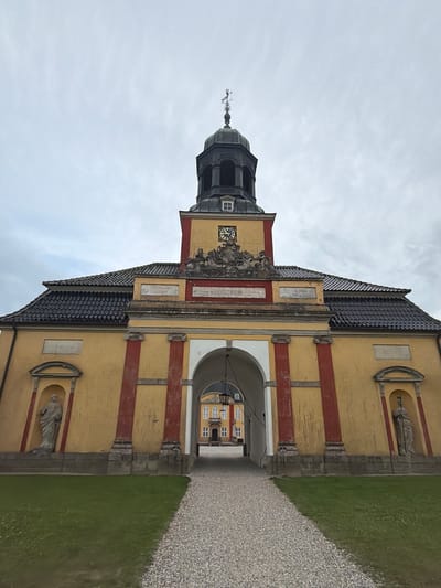 A wide-angle, low-angle shot depicts the exterior of a yellow building with a red-trimmed arched entryway, located in Lejre, Denmark. The building features a dark tiled roof and a central tower topped with a weather vane. On either side of the entryway, within arched niches, stand two stone statues. A gravel path leads towards the archway, with a strip of green grass to the left. Through the archway, another building with a yellow facade and white-trimmed windows is visible. The sky is overcast.