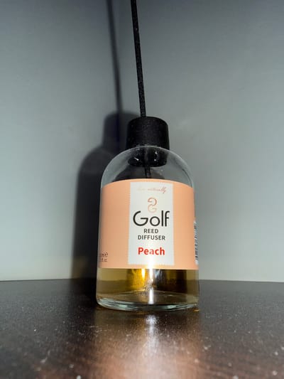 The image shows a close-up of a reed diffuser bottle lying on its side. The bottle is made of clear glass and contains a light amber-colored liquid. A black cap with a reed stick inserted into it is visible on the right side of the bottle. The bottle has a label with peach-colored text. The visible text on the label reads "Golf Reed Diffuser Peach" and "100 ml e 3.3 fl. oz.". There is also a small logo that appears to be a stylized "G". The background is dark and out of focus, with a lighter gray area to the right of the bottle. The lighting suggests an indoor setting. There are no people or animals in the image. The overall scene is a product shot of the reed diffuser. The image is oriented vertically.