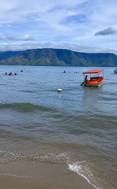 In Situngkir, Indonesia, a serene lake scene unfolds under a partly cloudy sky. The foreground features a sandy beach where gentle waves lap the shore, with white foam tracing the water's edge. The lake itself is calm, with a few white buoys dotting the surface. In the middle ground, several people are visible in the water, appearing to swim or float. Closer to the right side, a small, orange pedal boat with a canopy is moored. A person is seated in the boat, looking towards the left. In the background, a majestic mountain range with lush green slopes stretches across the horizon, its peaks softened by the distance and the haze. The water reflects the sky and the mountains, creating a tranquil and picturesque landscape.