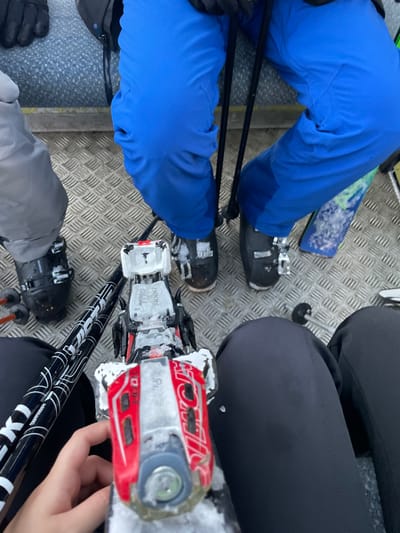 The image captures a high-angle view of multiple individuals and their ski equipment on a metal grated surface, likely within a ski lift or platform at a ski resort. The setting indicates cold, snowy conditions, with visible snow and ice clinging to the ground, ski boots, and bindings. Given the context, this scene is set in Courmayeur, Italy.

Several people are present, identifiable by their legs, boots, and a visible hand. On the left, a person wears vibrant blue ski pants and black ski boots, with black ski poles extending downwards. A portion of a ski with a blue and green pattern is visible behind their leg. In the mid-right, another person is wearing black ski pants. A hand, likely belonging to this person or another nearby, is seen holding the tip of a ski. At the bottom of the frame, a third person wears grey ski pants and black ski boots.

The ski equipment is a central focus. A prominent red and white ski binding, clearly branded "ATOMIC," is situated in the center-right. Adjacent to it is a white ski binding, partially obscured by snow and ice, with the text "DOOH" and "GULU PROME" visible. Black ski poles, potentially branded "LEKI" with white graphics, are crossed in the lower center. The metal grate flooring is typical of ski infrastructure, suggesting individuals are either waiting, taking a break, or in transit during a winter sports activity. The time of day appears to be daytime, with a somewhat subdued light, possibly overcast.