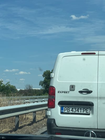 The image shows the rear of a white van with a Bulgarian license plate. The license plate reads "PB 4341 PE". The van appears to be parked on the side of a road, with a metal guardrail in the foreground. The sky is blue with some white clouds. The background shows trees and vegetation. The location is identified as Shtarkovo, Bulgaria.