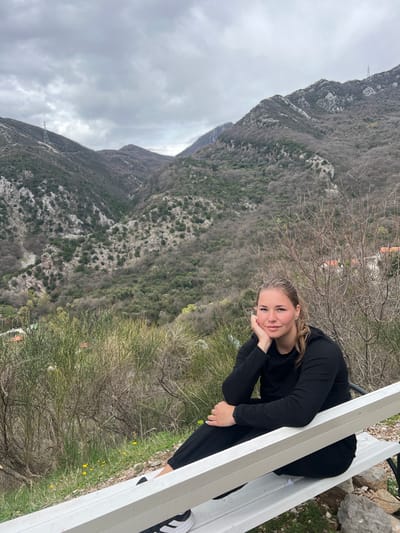 A young woman sits on a white bench, resting her head on her hand, gazing towards the camera. She wears a long-sleeved black shirt and black pants. Behind her, a mountain slopes steeply upward, covered with trees and shrubs. The sky is overcast. The landscape appears to be located near Krapina, Montenegro. In the foreground are a few small bushes.