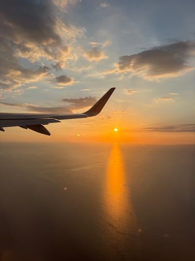 The image captures a view from an airplane window during sunset, with the right wing of the aircraft visible in the foreground. The sun is low on the horizon, casting a warm, orange glow across the sky and reflecting a shimmering path of light on the calm sea below. Scattered clouds, tinged with the sunset colors, dot the sky. The scene is serene and evocative of travel. The prompt incorrectly specifies the location as Isola Sacra, Italy; the image does not contain any identifiable landmarks or features that confirm this location.