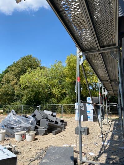 The image depicts a construction site in Hückelhoven, Germany. The scene is dominated by scaffolding, construction materials, and a natural backdrop of trees and a clear blue sky, suggesting it is daytime on a sunny day.
In the foreground and midground, various construction elements are visible. Scaffolding structures, made of metal, are prominent, with perforated metal walkways. Piles of dark grey, rectangular building blocks or insulation material are scattered on the ground, some neatly stacked and wrapped in plastic sheeting. Several white buckets, some with red branding, are also present. Electrical cables snake across the ground, indicating active work. A temporary metal barrier fence encloses part of the site.
Behind the construction elements, lush green trees with dense foliage create a natural boundary. The bright blue sky with a few wisps of white cloud suggests fair weather. The presence of construction materials and scaffolding clearly indicates ongoing construction or renovation work.
Visible text includes "Knauf" on some of the stacked materials. The branding on the buckets is partially obscured but appears to contain red lettering. The overall impression is one of a functional, active construction site set against a pleasant outdoor environment.