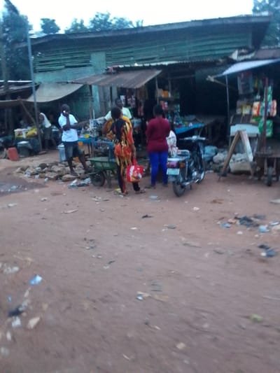 The image captures a bustling street scene, likely a market or commercial area, with an unpaved, reddish-brown dirt road in the foreground. Several people are visible, engaged in various activities.
In the center-left, a man in a white t-shirt and dark shorts stands, holding a phone to his ear. To his right, a person wearing a colorful patterned outfit (yellow, red, and black) stands next to a small green two-wheeled cart, holding a red bag. The word "RAM" is partially visible on the side of this cart. Further to the right, another person in a dark red top and blue trousers is seen near a parked motorcycle.
The background is lined with simple structures that appear to be shops or market stalls, characterized by their corrugated iron roofs. Goods are displayed, though indistinctly, on tables and shelves within these stalls. A blue table or stand is visible under one of the stalls. Rocks, debris, and scattered litter are present on the dirt ground, adding to the informal market atmosphere. Trees are visible in the distance, above the buildings.
The soft lighting suggests it might be late afternoon or early evening, or an overcast day. The scene depicts typical daily life in a semi-urban or rural setting, consistent with the provided location context of Orlu, Nigeria.