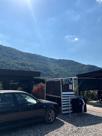 A black Volkswagen Golf is parked on a gravel lot in front of a building with a black facade. A small kiosk with blue and white stripes sells t-shirts. A sign on the kiosk reads "SUVENIRI DIVIC" with a heart symbol. In the background, a lush green mountain rises under a bright blue sky with a few scattered clouds. The scene appears to be in Divic, Bosnia and Herzegovina, given the sign.