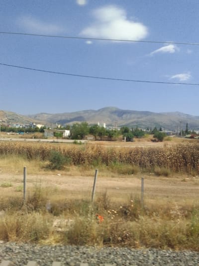 The image shows a view of a rural or suburban landscape with a clear blue sky and some scattered white clouds. In the foreground, there is dry vegetation and some wooden fence posts. In the middle ground, there are some buildings, including what appears to be a small white structure and some trees. Further in the background, there are hills or mountains, with some signs of habitation such as buildings and power lines visible on the slopes. The overall impression is of a dry, possibly Mediterranean environment. This scene is identified as being in the Municipal Unit of Ano Liosia, Greece.