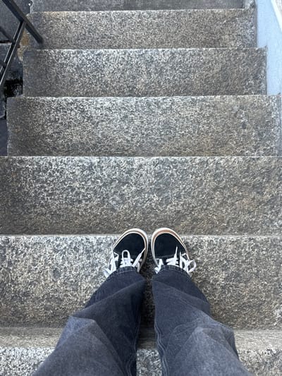 The scene captures a top-down view of an individual's lower body on a stone staircase in Visp, Switzerland. The person is wearing dark grey denim trousers and black canvas sneakers featuring white laces, white rubber soles, and a tan stripe accent. The feet are positioned on the second visible step from the foreground. The staircase consists of multiple steps constructed from coarse-grained, mottled grey stone, with at least six steps receding upwards from the viewer's perspective. A black metal handrail is visible along the left side of the stairs, extending into the lower left quadrant. A light-toned vertical surface borders the right side of the staircase in the upper portion of the frame.