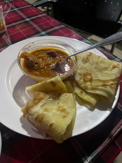 The image captures a close-up view of a meal served on a white round plate, which rests on a red and black plaid tablecloth. The plate holds several pieces of golden-brown flatbread, likely paratha, folded and showing some light browning, suggesting a soft and flaky texture. Adjacent to the flatbread, a clear glass bowl contains a rich, reddish-brown stew or curry with visible chunks of meat, possibly beef or goat, and a thick sauce. A silver metal spoon is placed within the stew.
The scene depicts a meal prepared for consumption. The dark background, coupled with the direct artificial lighting that casts shadows from the plate and food, suggests the photograph was taken in the evening or night, or in an indoor setting with low ambient light. A dark, indistinct object, possibly a piece of furniture, is partially visible in the upper left background. There are no people or visible text in the image. The food presentation is simple and functional, indicative of a casual dining experience.