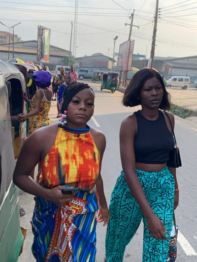 This image captures a daytime street scene in the city of Aba, Abia, Nigeria. In the foreground, two young Black females are prominent. The female on the left wears a multi-colored halter-neck top featuring orange, yellow, and blue patterns, paired with a blue and white patterned wrap or skirt. Her braided dark hair includes several light-colored beads on the left side. She holds a dark smartphone in her right hand and looks directly towards the camera with a neutral expression. The female on the right, positioned slightly behind the first, wears a black sleeveless crop top and loose-fitting teal-colored trousers with dark geometric patterns. She has short, dark, bob-length hair and carries a small black shoulder bag in her left hand, and another patterned bag in her right, while looking slightly to her right.
To the left, part of a green and black three-wheeled vehicle (rickshaw) is visible, with another female in a yellow patterned outfit and a purple head covering standing near it. The background displays a paved road occupied by multiple green three-wheeled rickshaws, a silver car/SUV, and a white van. Utility poles with extensive wiring, streetlights, and large commercial billboards are visible along the road. Distant buildings with light-colored facades and corrugated roofs line the street. The sky is a light blue with minimal cloud cover.