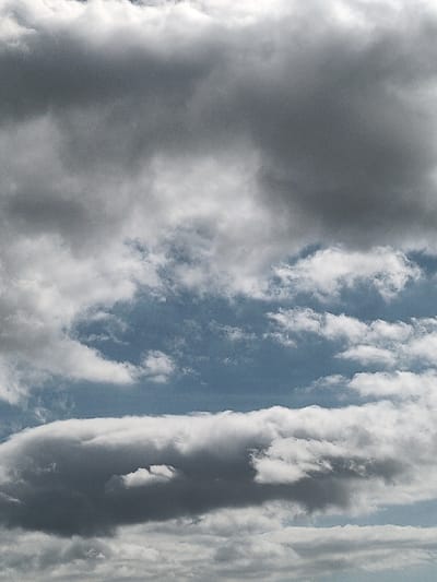 The image displays a sky scene over Ojos de Garza, Spain. The frame is predominantly filled with a mix of cumulus and stratocumulus clouds. The upper section features dense, dark grey cloud formations, indicative of significant thickness. Below these, lighter white and grey clouds are interspersed with clear patches of medium blue sky, particularly evident in the mid-horizontal plane. A distinct, elongated dark grey cloud with a brighter rim occupies the lower-mid portion of the frame, underneath which additional layers of white and light grey clouds are visible. There are no identifiable people, man-made subjects, objects, actions, or interactions present in the depicted environment, which consists solely of atmospheric elements.
