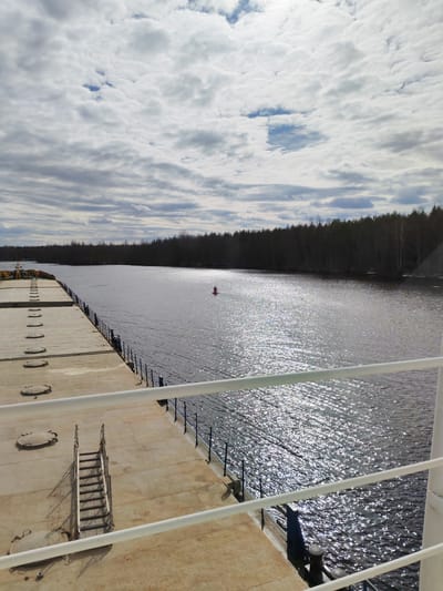 The image captures a wide inland waterway, likely a river or canal, viewed from the deck of a large vessel, possibly a cargo ship or barge. In the foreground, the ship's expansive, flat deck is visible, featuring several oval-shaped hatch covers and a portable metal staircase. White railings run along the side of the deck, partially framing the scene.
Beyond the ship, the dark water shimmers intensely with sunlight reflections, suggesting a bright day despite the cloudy sky. A red, conical navigational buoy floats in the mid-distance on the water, indicating a marker in the channel. The far bank of the waterway is lined with a dense, dark forest of deciduous trees, mostly bare, implying either early spring or late autumn. The tree line extends far into the background, following the course of the water.
The sky above is predominantly covered with light grey and white stratocumulus clouds, with occasional patches of blue sky visible through breaks in the cloud cover. The overall atmosphere is calm and expansive. No people or visible text are present in the image. The scene suggests a vessel navigating or moored in a natural or semi-natural environment, characteristic of an inland waterway in Kurdiug, Russia.