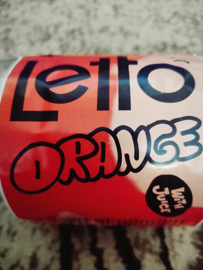 The image shows a close-up of a beverage can. The can is primarily red and white with black text. The word "Letto" is written in large black block letters across the top of the can. Below this, in a bubbly, graffiti-style font, is the word "ORANGE". To the bottom right, on a black circular sticker, are the words "WITH JUICE" written vertically in white. The background appears to be a textured surface, possibly a carpet or fabric, with muted colors. The lighting is somewhat dim, and there is a noticeable graininess to the image, suggesting it was taken indoors under artificial light or with a low-quality camera. No people are visible. The image focuses entirely on the product packaging. Based on the provided location context, this image was likely taken in Vatra, Moldova.