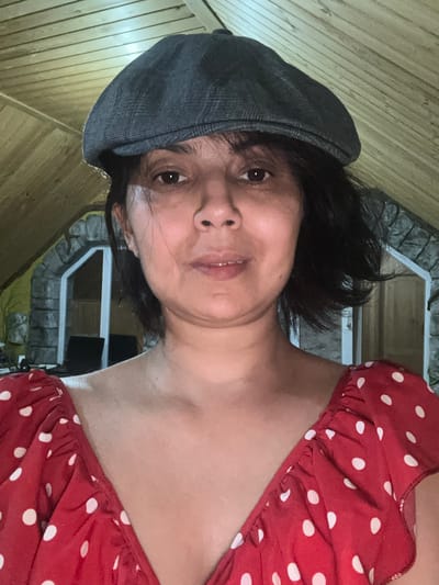 A woman is looking directly at the camera, wearing a grey flat cap and a red and white polka dot dress. The background shows a sloped wooden ceiling and a stone arched doorway. The image is a close-up, head and shoulders shot, taken indoors, likely in an attic or loft space. The overall lighting is dim, with a slight warm tone. There are no identifiable landmarks that would place this image specifically in Kalininskaya, Russia.