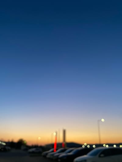 This is a vertical, slightly blurry, eye-level shot of a sunset over what appears to be a parking lot in Bolesławiec, Poland. The sky transitions from a deep, dark blue on the left side of the frame to a soft orange and yellow gradient on the right, indicating the setting sun. In the background, on the right side of the image, there are several blurred vertical objects that appear to be streetlights or possibly buildings. A line of blurred, dark-colored cars is also visible parked along a road. The overall mood is peaceful and calm, with the soft colors of the sunset creating a serene atmosphere. There is no visible text in the image. The time of day is clearly dusk or twilight.