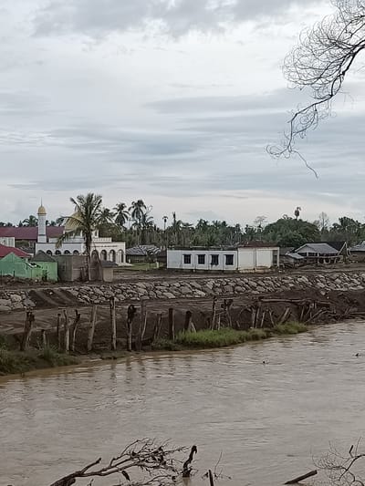 The image captures a scene in Meunasah Raya, Indonesia, featuring a river in the foreground with muddy, brown water. Along the riverbank, a damaged wooden fence constructed from vertical posts and horizontal beams is visible. Beyond the fence, there is a rocky embankment and then a collection of buildings. Prominently, a mosque with a golden dome and a white minaret stands out, surrounded by other structures including smaller, less elaborate buildings and what appear to be houses. Vegetation, including palm trees and denser foliage, can be seen in the background, suggesting a tropical or sub-tropical environment. The sky is overcast with grey clouds, indicating a cloudy or potentially rainy day. The overall impression is one of a developing or possibly affected community situated near a natural water source. There are no people clearly visible in the image.