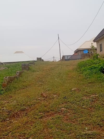 The image depicts an outdoor scene in Ashata, Nigeria, featuring a sloped, grassy terrain with scattered rocks and some remnants of concrete structures. In the background, several buildings are visible. To the left, a large, white, gabled roof structure is prominent. Further to the right, a light-colored building with a dark tiled roof is situated, and next to it, another building partially obscured by foliage, featuring a blue satellite dish mounted on its roof. A metal gate is also visible between the buildings. A wooden utility pole with electrical wires stretches across the sky. The overall atmosphere is overcast, suggesting cloudy weather. There are no people visible in the image.