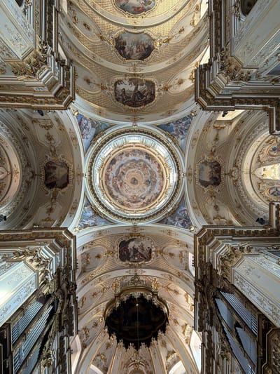 The image is a low-angle shot looking up at the ornate ceiling and dome of a church, likely within the historic center of Mozzo, Italy. The architecture features a white and gold color palette with intricate detailing. The ceiling is adorned with numerous frescoes depicting religious scenes and figures, framed by elaborate gold trim and decorative patterns. A large, central dome with its own frescoed interior is visible, illuminated by natural light filtering from above. Flanking the central dome are smaller arched sections also decorated with frescoes. In the foreground, the pipes of a large pipe organ are visible, with their metallic sheen contrasting with the opulent gold decorations surrounding them. The overall impression is one of grandeur and religious artistry.