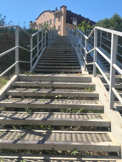 The image depicts an outdoor metal staircase with white railings, ascending a grassy slope under a clear blue sky. The staircase is made of metal grating for the steps and solid concrete for the risers. Some green vegetation is growing through the metal grating of the steps. In the background, a multi-story brick building with a dark roof is visible, surrounded by trees. The location is identified as Verkhnetemernitsky, Russia. There are no people or animals present in the image. The overall scene suggests an urban or suburban environment with public infrastructure.