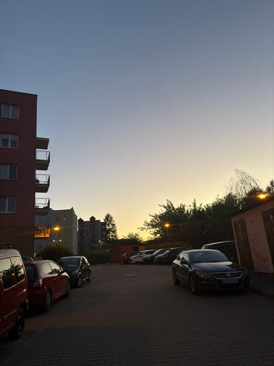 The image depicts a residential area in Lublin, Poland, at dusk. A multi-story apartment building, painted a reddish-pink, dominates the left side of the frame, its balconies illuminated by the fading sunlight. Across from it, other apartment buildings of varying heights and colors are visible, with streetlights casting a warm glow on the pavement. A parking lot fills the foreground and middle ground, with several cars parked, including a dark Volkswagen Passat with license plate LUB 90162. Trees and bushes line the edge of the parking lot, and a small utility building with graffiti is visible on the right. The sky is a gradient of pale blue to soft yellow, indicating the setting sun. The scene is quiet and still, with no people visible.