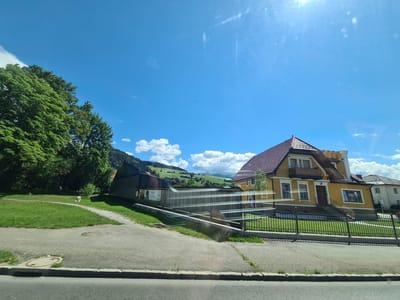 The image shows a residential area on a sunny day with blue skies and scattered clouds. In the foreground, there is a paved road with a curb and a manhole cover. To the left of the road is a grassy area with a path winding through it. Several trees with lush green foliage are on the left side of the image, and a small, light-colored animal, possibly a dog, is visible on the grass near the trees.

In the middle ground, there are several buildings. A long, low building with a dark roof and wooden siding is prominent. Adjacent to it is a larger, two-story house painted yellow with a reddish-brown tiled roof. This house has multiple windows, a balcony on the upper floor, and a dark wooden door with a set of stairs leading up to it. A black metal fence runs along the front of the yellow house and a portion of the darker building.

In the background, rolling green hills with some scattered houses and trees can be seen, suggesting a semi-rural or suburban setting within the city of Sankt Marein bei Neumarkt, Austria. The sun is shining brightly, casting a slight glare on the image, particularly in the upper right corner. The overall scene conveys a peaceful, pleasant day in a scenic Austrian location.