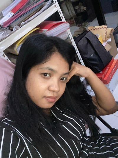 The image shows a person lying on a bed. The person has long black hair and is wearing a black and white striped shirt. The person's right hand is placed near their forehead. The background includes a shelf with documents and other items. There are packages, a box, and a black bag on the shelf. The location is Negara, Indonesia.