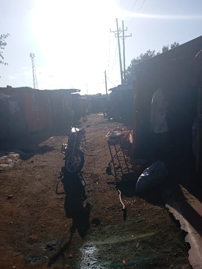 The image depicts a bright, sunlit alleyway or unpaved street, likely in a residential or commercial area of a town like Namanga, Kenya. The scene is characterized by small, informal structures lining both sides of the dirt path, many appearing to be constructed from corrugated metal or similar materials.
In the foreground on the left, a motorcycle, commonly known as a boda-boda, is parked on the dusty ground. To its right, closer to the center, is a metal stand or table. Further along the right side of the path, inside the entrance of one of the structures, a light-colored garment is seen hanging. Near the metal stand, a large, dark bag rests on the ground, alongside a long stick or branch. The ground itself is uneven, with patches of mud and dry earth, reflecting the bright sunlight in some areas.
The harsh, direct sunlight from the top left of the frame casts long, distinct shadows, most notably the shadow of a person (presumably the photographer) extending from the bottom center towards the motorcycle. This strong lighting suggests it is late morning or midday under a clear sky.
In the background, utility poles with overhead power lines rise prominently on the right side of the path, stretching into the distance. On the far left, a tall communication tower, likely a cell tower, is visible against the bright sky.
Visible text includes the word "Airtel" prominently displayed in white letters on a red background on the side of one of the structures on the left, with smaller, less legible text (possibly "Money") beneath it. The overall impression is one of everyday life in a bustling, yet quiet at this moment, local setting.