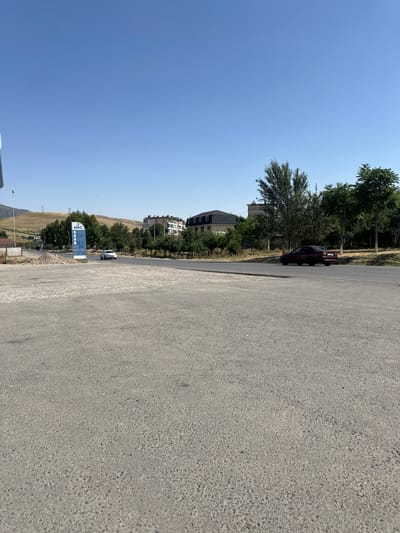 The image depicts a roadside scene in Розиён, Tajikistan. A paved road runs horizontally across the middle ground, with a white sedan driving on it from left to right. To the right of the road, a dark red sedan is parked or moving slowly. In the background, there are buildings, trees, and a hilly landscape under a clear blue sky. A blue and white signpost displaying prices, likely for fuel, is visible on the left side of the road. The foreground consists of a rough, grey paved area.