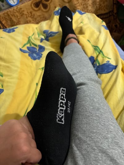 The image shows a person's legs and feet lying on a bed with a yellow floral patterned sheet. The person is wearing black low-cut socks with the brand name "Kappa" and the size "39/42" visible on the right sock. They are also wearing grey sweatpants. A brown patterned blanket is visible at the top of the image, and a blue alarm clock is partially visible on the left. The scene appears to be indoors, in a residential setting. There is no discernible information in the image that indicates a specific location in the city of Zgorigrad, Bulgaria.