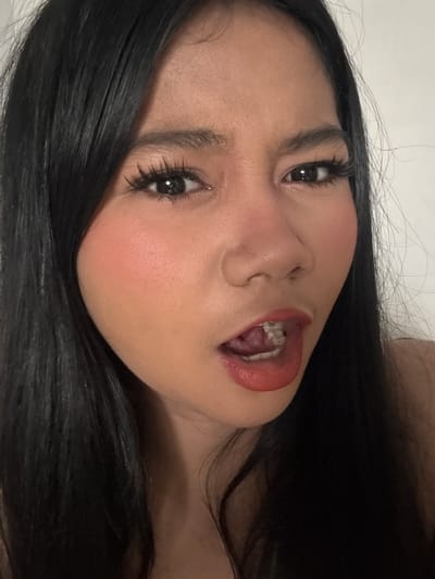 A close-up, headshot of a young woman is depicted. She has long, dark hair framing her face, which is turned slightly to the side. Her eyes are wide open, with prominent eyelashes, and her eyebrows are neatly shaped. Her nose is slightly tilted, and her lips are parted, revealing her teeth. She is wearing coral-colored lipstick and blush. The background is a plain, light-colored wall. This image was taken in North Jakarta, Indonesia.