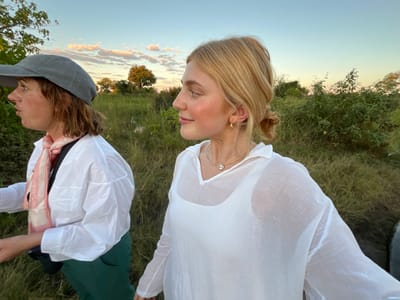 Two women stand outdoors in Lobilos, Botswana, surrounded by grassy terrain with scattered trees and shrubs under a blue and light orange sky. The woman on the right has blonde hair pulled back, wears gold hoop earrings, a gold necklace, and a white sheer long-sleeved shirt over a white tank top. She is looking to the right of the frame. The woman on the left wears a gray baseball cap, a white long-sleeved shirt, green pants, and a light pink and white scarf. A black object is strapped across her chest. The area appears to be grassy with green vegetation and sparse trees in the background.