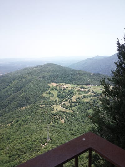 An aerial, long shot captures a verdant, mountainous landscape under a hazy sky. The foreground features a dark brown metal railing, partially obscuring the view. Below the railing, a dense forest covers the rolling hills. In the mid-ground, a small village with red-roofed buildings is nestled in a clearing, surrounded by fields and trees. A winding dirt road snakes through the landscape. In the background, layers of misty blue mountains recede into the distance. A tall, lattice-like power line tower stands on a hillside, its wires disappearing into the trees. The overall scene depicts a rural, mountainous region, likely in Susqueda, Spain, known for its natural beauty and sparse settlements.