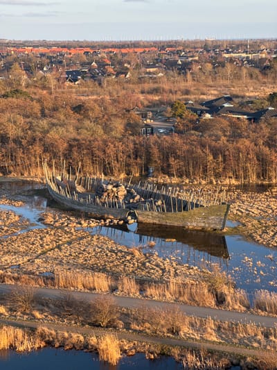 This image captures an aerial, panoramic view of a wetland landscape transitioning into a suburban and urban environment within Tårnby, Denmark, under natural lighting conditions.

In the mid-ground, a large, weathered wooden boat, appearing skeletal in structure, is partially submerged in a shallow body of water surrounded by dense, dry, golden-brown reeds. Numerous slender wooden stakes are vertically embedded around and within the boat's hull. A prominent pile of natural, rounded stones and rocks rests within the central section of the boat. The water immediately surrounding the boat reflects the sky and portions of the structure.

The immediate foreground features a narrow, unpaved or dirt path running horizontally, bordered on both sides by tall, dry reeds and sparse, leafless bushes. Parallel to this path, a darker, calmer body of water is visible in the lower foreground.

Beyond the boat structure, extending towards the mid-background, is a dense expanse of deciduous trees, mostly bare of leaves, displaying shades of brown and reddish-orange, consistent with late autumn or winter. Scattered within this wooded area and further back are residential houses, distinguishable by their dark roofs and varying architectural styles.

The distant background reveals a more densely populated urban or town area characterized by numerous buildings, many of which have distinctive orange or red-tiled roofs. A prominent feature on the far horizon is a long, continuous line of numerous tall wind turbines, silhouetted against a light blue sky with faint cloud cover. No individuals are present in the scene. The overall lighting suggests either early morning or late afternoon, casting warm tones on the dried vegetation.